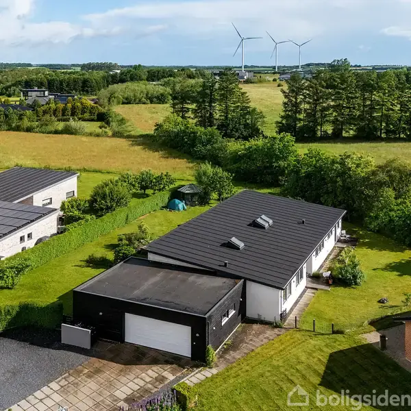 En moderne, sort-tagget bungalow med garage omgivet af frodige grønne områder og nabohuse, beliggende i et landligt område med vindmøller i baggrunden under en lys himmel.