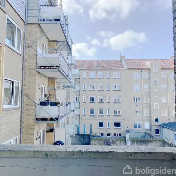 Lejlighedsbygninger står stille i en gård, fyldt med åbne vinduer og altaner. Bygningerne er opført i lyse mursten og omgivelserne domineres af en blå himmel med lette skyer.