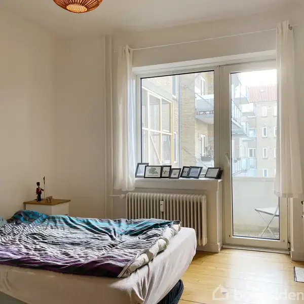 En seng er opredt ved siden af et natbord i et lyst værelse. Vinduet viser en altan og nabobygninger. Rammer står på radiatoren.