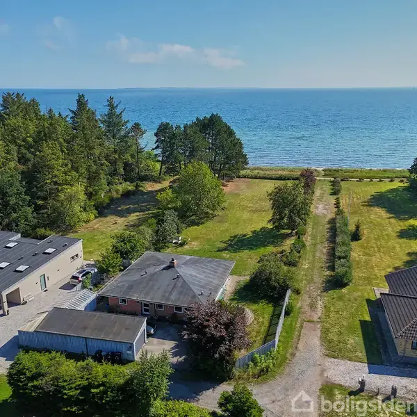 Et hus og en garage står stille ved siden af en grusvej. Omkring dem er der grøn vegetation. I baggrunden ses en strand og det åbne hav under en blå himmel.