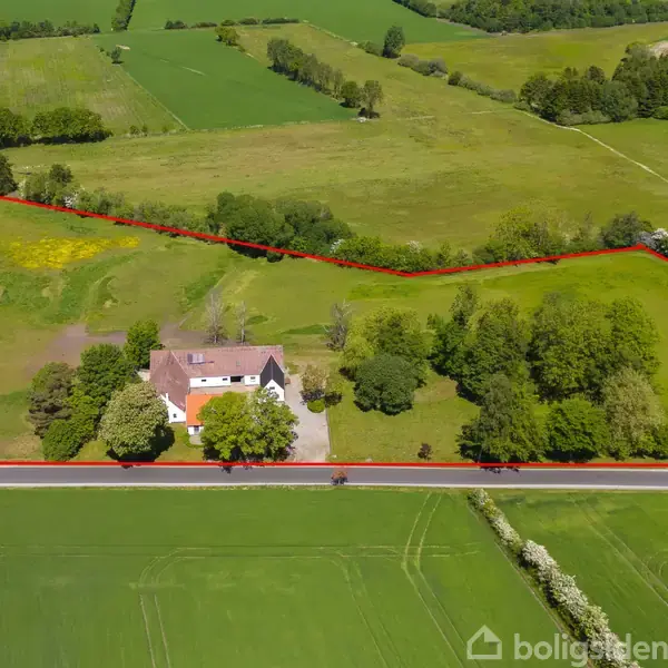 Hus omgivet af træer; ligger på en rummelig grund afgrænset med rød linje. Vejen løber foran huset, og omkringliggende marker strækker sig ud i baggrunden. Tekst: "Boligsiden".