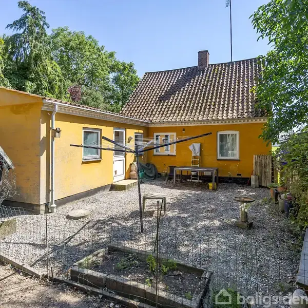 Et gult hus med rødt tegltag står i en gårdsplads med grus. Der er et tørrestativ og et lille hegn, omgivet af træer og buske.