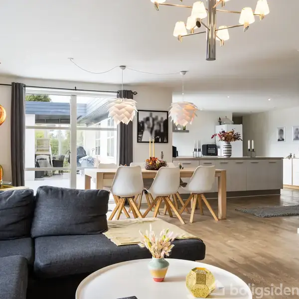 En moderne stue med en grå sofa, sofabord og lysende loftlamper. I baggrunden ses en spisestue med seks stole og et åbent køkken. Store vinduer giver naturligt lys.