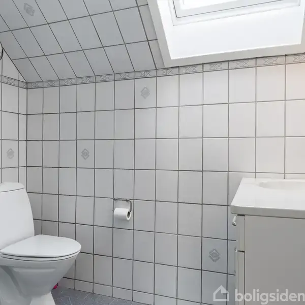 Toilet hvilende under skrå loft, omgivet af hvide fliser med mønster. Ved siden af er et hvidt badeværelsesskab med vask, over hvilket der er et spejl og en tandbørste i holder.