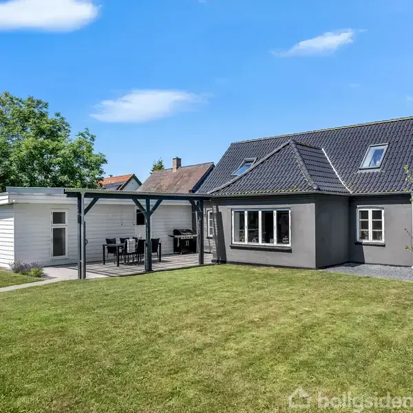 Et gråt hus med sort tag står på en velplejet græsplæne. Ved siden af er en terrasse med havemøbler under et halvtag. Træer omkranser haven.