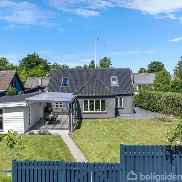 Et hus med sort tag og hvide vinduer står i en grøn have med en trampolin. En blå himmel og flere træer omkranser området.