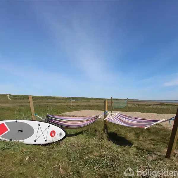 Et stand up paddleboard ligger på græs ved siden af to hængekøjer spændt mellem stolper. De befinder sig i et åbent naturområde med grønne marker og blå himmel.