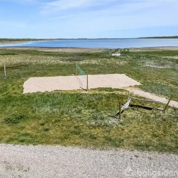 Volleyballbane med net står ubenyttet på græsset ved siden af en sø med åbent landskab i baggrunden. Stier og frodig natur omgiver banen.