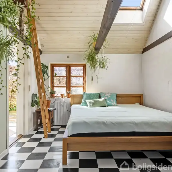 Et soveværelse med en stor seng står under et skråloft med ovenlysvinduer. Planter hænger fra loftet og en stige fører til en hems. Rummet har sort-hvidt ternet gulv.