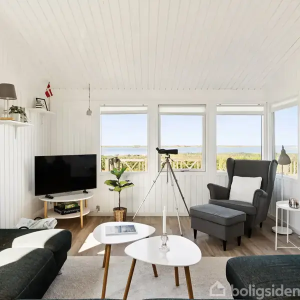 En lys, moderne stue med hvide vægge og store vinduer. En sofa og lænestol er placeret omkring et sofabord. En kikkert står ved vinduerne, med en udsigt over naturen udenfor. En fladskærms-tv er på en reol.