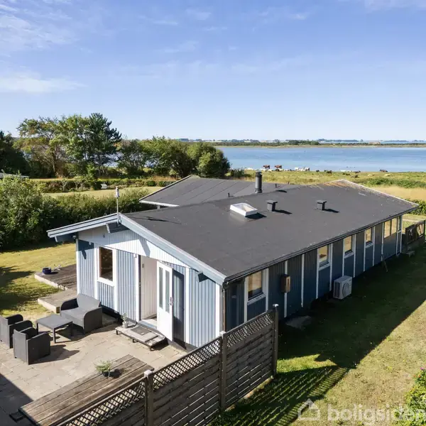 Et blåmalet træhus står stille ved en terrasse med havemøbler. Bag huset ses en smuk udsigt over vandet, omkranset af grønne buske og træer.