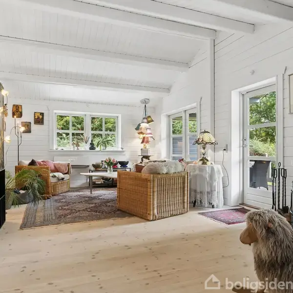 Stue med træmøbler og tæpper; store vinduer og en åben dør til verandaen. Rummet er lyst med hvidmalede vægge og synlige bjælker, dekoreret med planter og lamper.