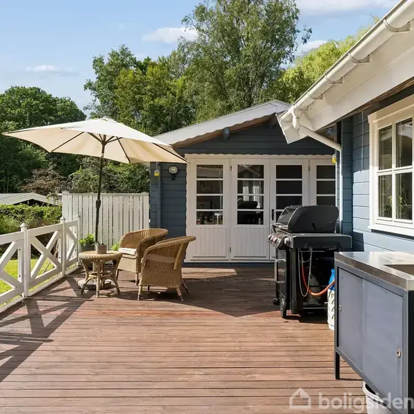 Træterrasse med et udendørs køkken og grill står ved et blåt hus. To kurvestole og et lille bord er placeret under en parasol. Grønne træer omgiver området.