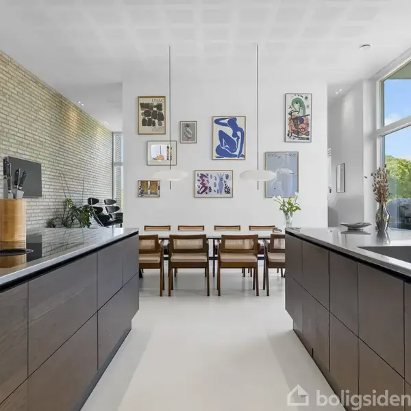 Et moderne køkken-alrum med to øer, æstetiske træskabe, og spisebord omgivet af stole. Væggen har flere kunstværker, og store vinduer giver udsigt til det grønne udenfor.