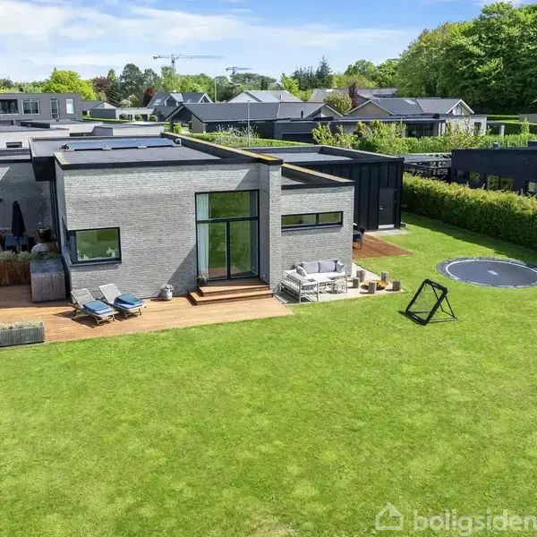 En moderne villa med fladt tag står på en stor, grøn græsplæne, omgivet af hække. Der er terrasse med loungemøbler, liggestole og en trampolin i haven.