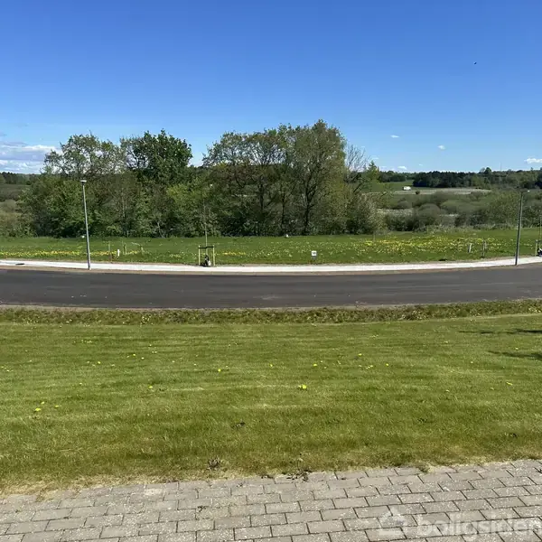 En nylagt asfaltvej skærer gennem grønne områder med træer i baggrunden under en blå himmel. Græsplænen i forgrunden er velplejet og omkranser en flisebelagt sti.