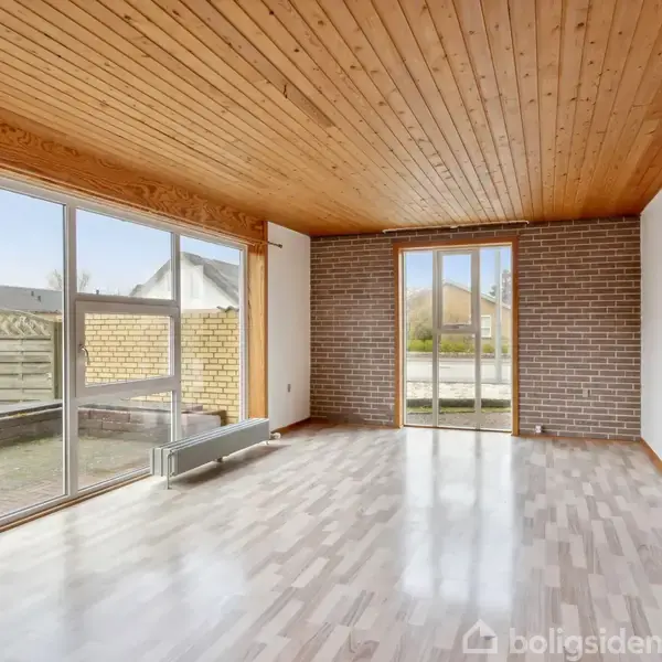 Stue med trægulv og træloft, store vinduer og glasdør giver lys adgang til terrasse. En murstensvæg komplementerer det enkle interiør. Ingen tekst er fundet.