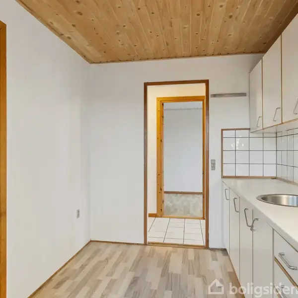 Køkken med hvide skabe og en vask på en træbordplade. Trægulv og træloft sammen med hvide fliser på væggen. En åben dør fører til en anden del af huset.