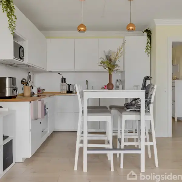 Køkkenet har hvide skabe og træbordplade, mikroovn, kaffemaskine, og høj bord med tre skamler. Grønne planter og vaser med tørrede blomster dekorerer rummet.