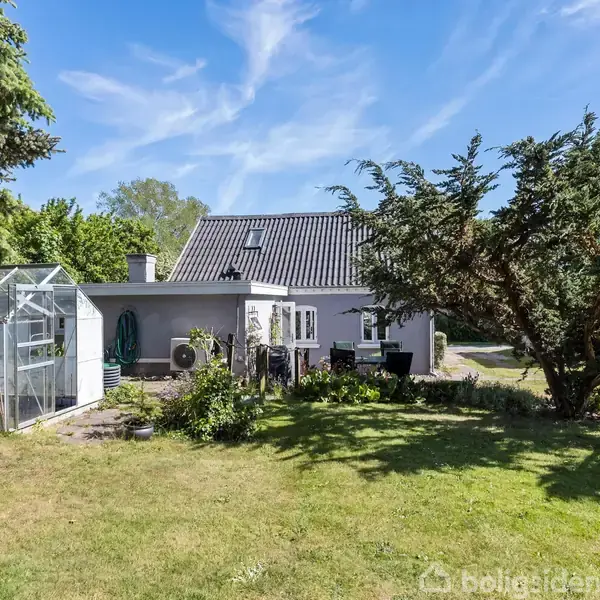 Et hus med en grå facade står i en have med græsplæne. Et drivhus er placeret til venstre. Store træer og blå himmel omkranser stedet.