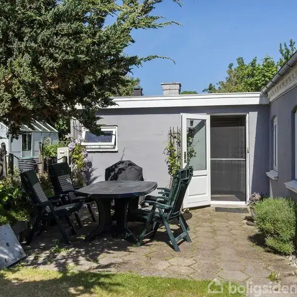 Et rundt bord med seks sorte stole står på en terrasse med fliser. Bordet er i skyggen af et træ, mens huset har lysegrå vægge. Græsplæne og have omkranser området.