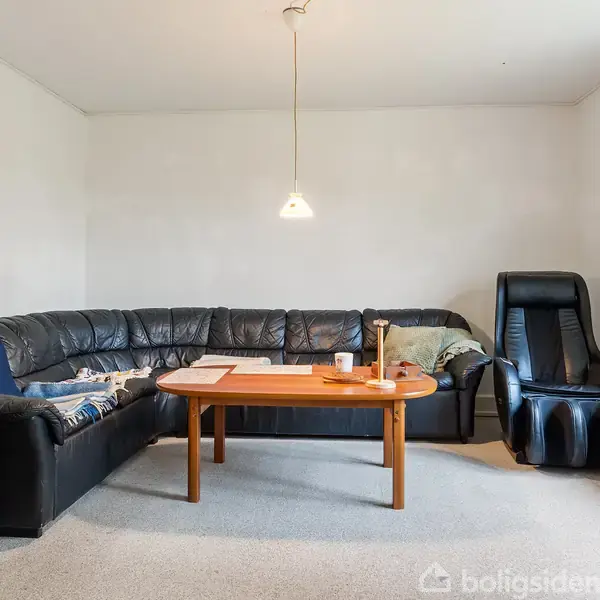 Sort hjørnesofa med tæppe og puder står i en stue ved siden af en lænestol. Et træ sofabord har magasiner og en kop. Lyse vægge og hængende lampe.