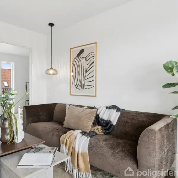 Sofaen er brun og placeret i en moderne stue med hvide vægge. Tæpper og puder dekorerer sofaen. En potteplante står ved siden af, og en indrammet kunstplakat hænger på væggen over sofaen.