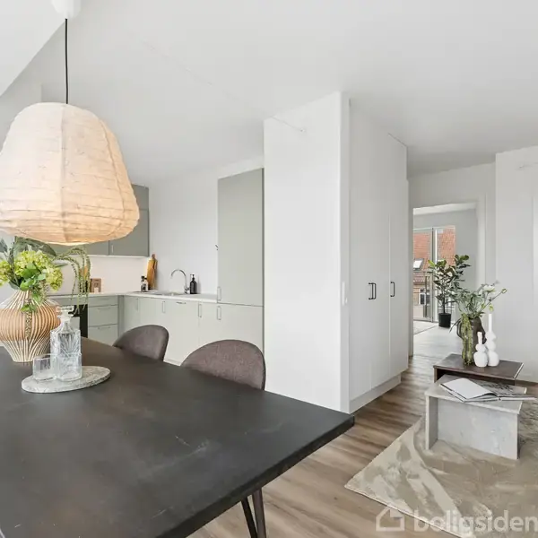 Spisebord med sort overflade og stole står i en lys stue med en stor lampe over. Til venstre ses et moderne køkken, og baggrunden viser stue med sofa og sofabord.