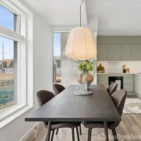 Et spisebord med brune stole står i et moderne køkken med grønne skabe. En stor lampe hænger over bordet, der er dekoreret med en vase med blomster. Store vinduer giver udsigt til tagene udenfor.