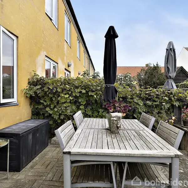 En træhavebord med stole står på en gårdhave omgivet af hække og en gul bygning. Paraplyer er placeret i bordets midte, og der er planter omkring.
