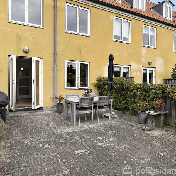En gul murstensbygning med flere vinduer har en flisebelagt terrasse foran. Terrassen har et træbord med stole, en bænk, planter og en grill, omkranset af lave trægærder.