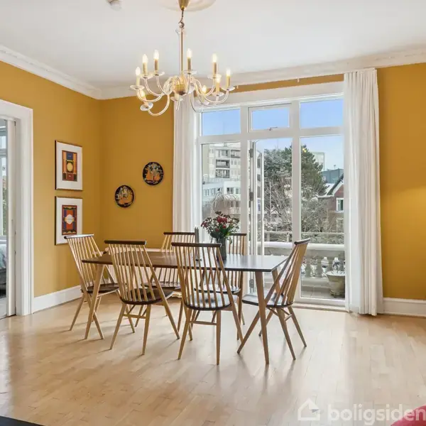 Spisebord med træstole står i en spisestue med gul vægfarve. En lysekrone hænger over bordet. Store vinduer viser udsigt til byen. En bænk er synlig i sidstuen.