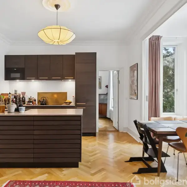 Køkken med mørke skabe og en ø, omgivet af lyst træparketgulv. Spisestue med vinduer og udsigt til træer. Planter og kunst pryder rummet.