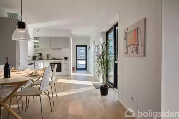 Et moderne køkken-alrum med spisebord og stole, i en lys og minimalistisk indretning. Der er en stor glasdør til højre og vægkunst ved siden af en potteplante.