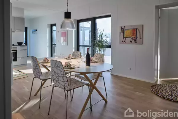 Et rundt spisebord er dækket med glas og en vinflaske i et lyst køkken-alrum. Fire moderne stole omgiver bordet, og en plante står ved vinduet. Vægkunst hænger på væggen.