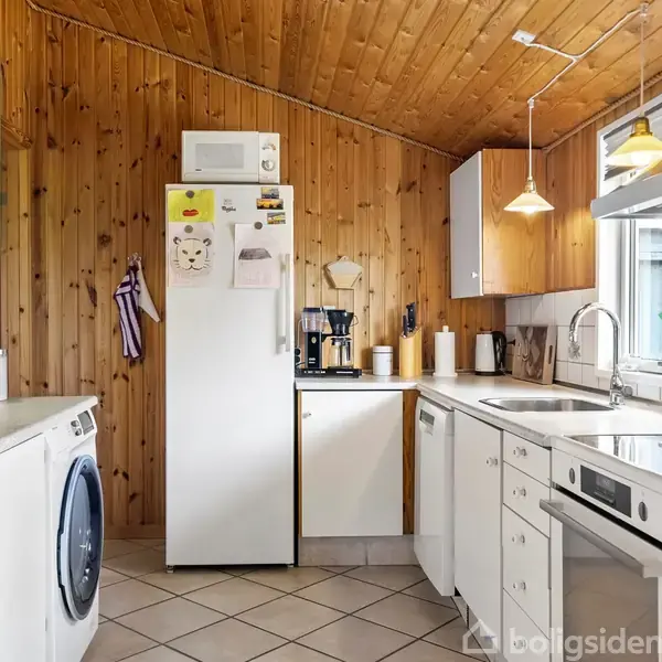 Et hvidt køleskab står i et træbeklædt køkken ved siden af en opvaskemaskine og vaskemaskine. Loftet er også af træ, og der hænger lamper over stålbordpladen.