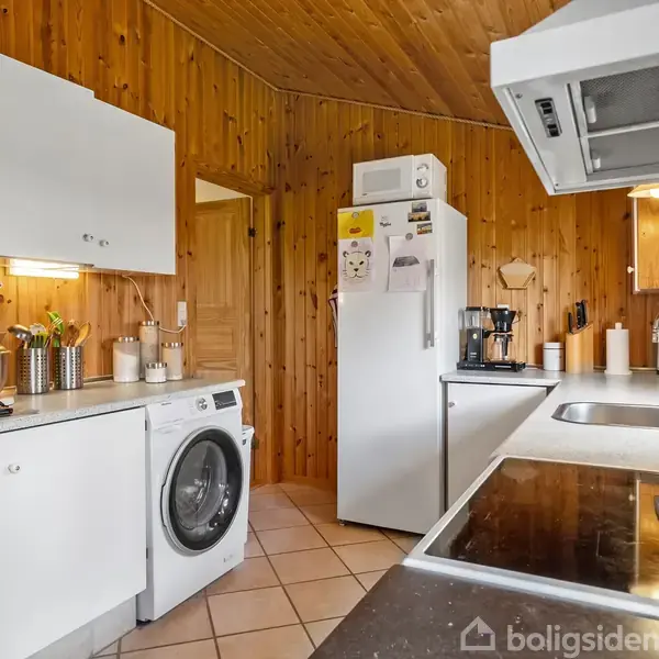 Køleskab står i køkken med trævægge og flisegulv, omgivet af vaskemaskine, kaffemaskine, komfur, og vask. Flere køkkenredskaber og krydderiglas ses på bordpladerne under overskabe.