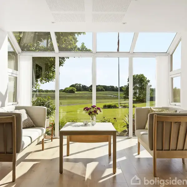 To sofaer og et bord med blomster står i en lys stue med store vinduer, med udsigt til en grøn mark og træer udenfor.