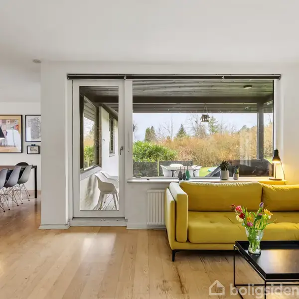 Gul sofa står i en stue mod glasparti med udsigt til have, placeret ved et sort sofabord med blomster. Til venstre er der spisebord med sorte stole og lamper.