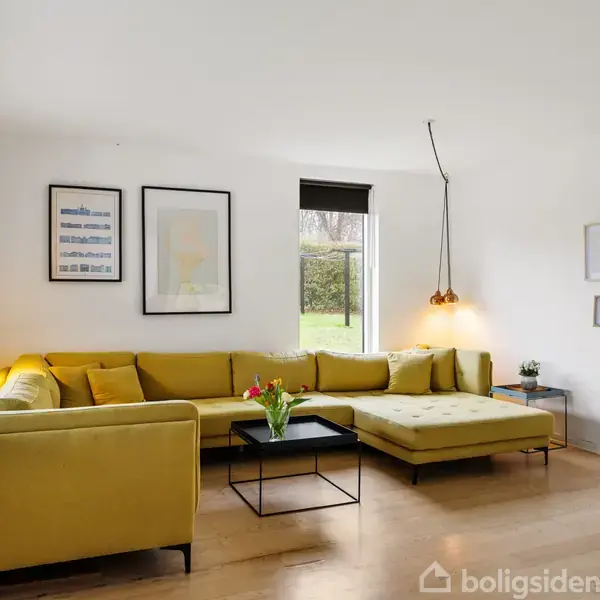 En gul sofa står langs væggen i en lys stue. På væggene hænger kunstværker, og gennem vinduet ses en have. En sort lampe hænger over sofabordet.