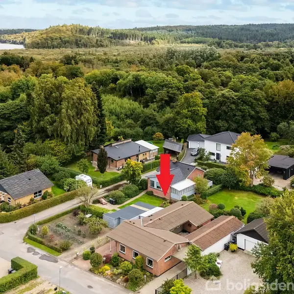Et parcelhus er markeret med en rød pil midt i et frodigt, skovbevokset område med flere omkringliggende huse. I baggrunden ses et kuperet terræn og en flod.