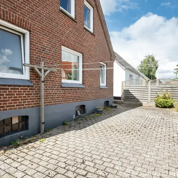 Et rødt murstenshus med vinduer står ved en belagt gårdhave. Et tørrestativ er fastgjort til husets væg. En træhegn omkranser området, og himlen er skyet.