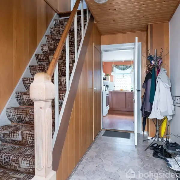 En trappe beklædt med mønstret tæppe fører opad. I gangen hænger jakker på en knagerække. Der ses også et rundt spejl og en dør til et køkken.