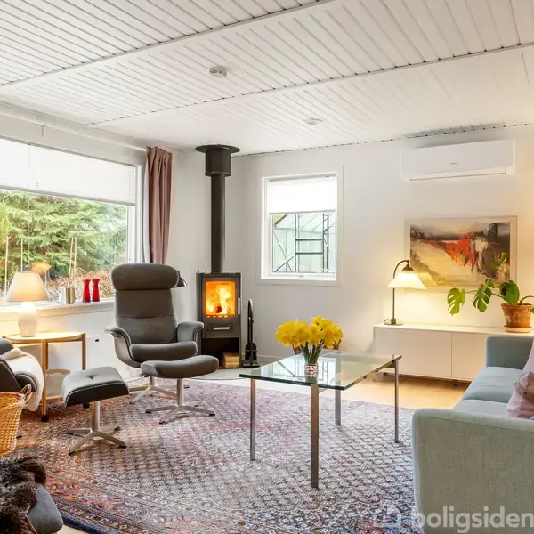 En hyggelig stue med brændeovn, der er i gang. Møblerne inkluderer en sofagruppe og lænestole. Store vinduer giver udsigt til naturen udenfor, mens lamper oplyser rummet indendørs. En vase med gule blomster står på et sofabord.