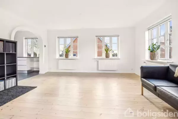 Stue med trægulv, sofa til højre og bogreol til venstre. Tre vinduer med planter giver udsigt til røde murstensbygninger. Lysindfald fra flere sider, tilstødende køkken.