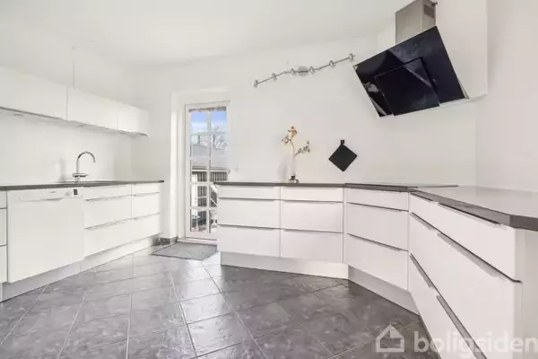 Et moderne køkken med hvide skabe og grå flisegulve, har en sort emhætte. I baggrunden ses en glasdør, der fører ud til en terrasse.