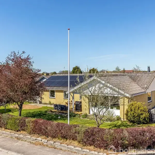 En gul murstenshus med solpaneler på taget står i en velplejet have med en flagstang. Huset er omgivet af buske og træer og ligger i et villakvarter.