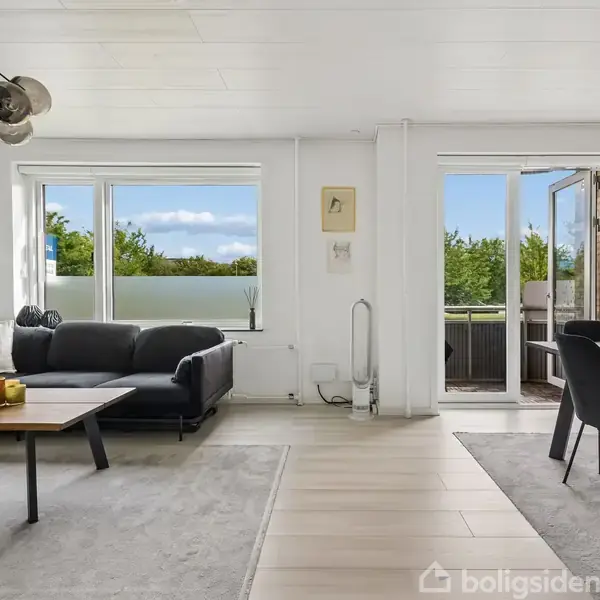 Stue med sort sofa og sofabord til venstre, en spisestue med seks stole til højre, lyse vægge og store vinduer med havedør, der fører ud til en terrasse.