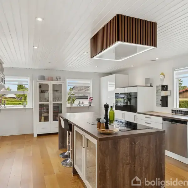 Et moderne køkken med en stor køkkenø og høje barstole. Sollys strømmer ind gennem vinduer, og der er en lampe med trædesignelementer over øen. Planter pynter rummet.