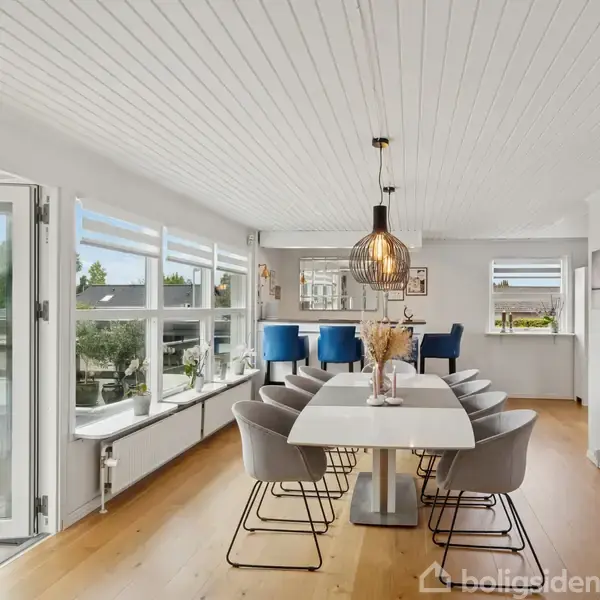 Langt spisebord med polstrede stole omgivet af store vinduer, der giver udsigt til en have. Moderne loftlampe hænger over bordet i et lyst og rummeligt spisestuemiljø.